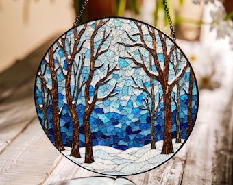 Op de afbeelding: Een rond glas-in-loodraam dat aan een ketting hangt. Het raam toont een winterscène met kale bomen en een besneeuwd landschap in tinten blauw en wit.