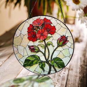 Puede incluir: Arte de ventana de vidriera redonda con un vibrante diseño de flores de geranio rojo. La obra de arte incluye hojas y capullos verdes, sobre un fondo texturizado blanquecino. Se adjunta una cadena de metal negro para colgar.
