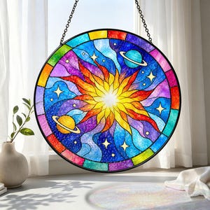 Puede incluir: Un atrapasoles circular de vidrieras con un sol central amarillo y naranja rodeado de patrones cósmicos arremolinados en azul y violeta, dos planetas con anillos y estrellas blancas, todo dentro de un marco negro con cadena.