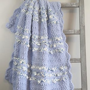 May include: A soft, light blue crochet blanket with white and light blue accents. The blanket has a scalloped edge and is draped over a wooden ladder. The texture appears plush and cozy, perfect for adding warmth and style to any space.
