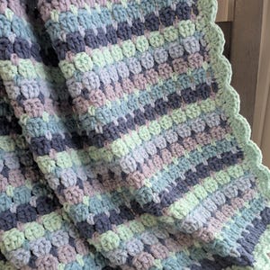 May include: A hand-crocheted blanket in various shades of blue, lavender, and mint green. The blanket has a textured stitch design and a scalloped border. The Clever Stitch Co. logo is visible in the bottom right corner.