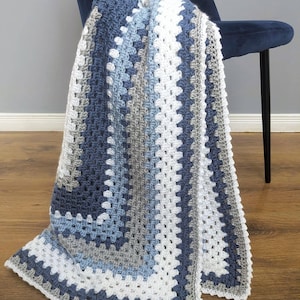 May include: A handmade crochet blanket in shades of blue, gray, and white, draped over a dark blue chair. The blanket features a granny square design with a textured appearance. The Clever Stitch Co. logo is visible in the bottom right corner.