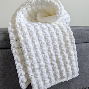 May include: A white, textured crochet blanket with a rolled-up section. The blanket has a repeating pattern of raised stitches, creating a soft, plush appearance. The blanket is draped over a gray surface, with the Clever Stitch Co. logo visible in the bottom right corner.