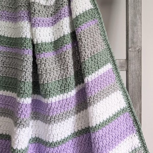 May include: A handmade crochet blanket with a striped pattern. The blanket features alternating stripes of white, gray, lavender, and sage green. The blanket is draped over a wooden ladder, showcasing its texture and design.