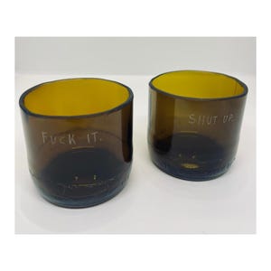 Könnte beinhalten: Zwei bernsteinfarbene Trinkgläser aus wiederverwerteten Flaschen. Ein Glas trägt die weiß gravierte Aufschrift "FUCK IT." und das andere Glas die weiß gravierte Aufschrift "SHUT UP.". Beide Gläser haben eine glatte, zylindrische Form.