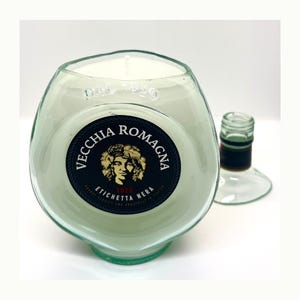 Op de afbeelding: Een kaars gemaakt van een hergebruikte wijnfles, gevuld met witte was. De kaars heeft een zwart etiket met gouden letters en een portret. De basis van de fles is groen en de tekst "VECCHIA ROMAGNA" is zichtbaar.