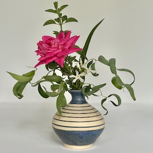 May include: A small, round ceramic vase with horizontal cream and navy blue stripes, holding a vibrant arrangement of a large pink rose, white flowers, and green foliage. The vase has a dark blue rim and base.