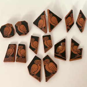 May include: A collection of terracotta-colored owl-themed earrings and pendants with black backgrounds. The owl designs are in a classical Greek style, with various geometric shapes and sizes. The jewelry pieces are arranged on a white surface.