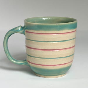 Puede incluir: Una taza de cerámica con asa y borde turquesa, y rayas horizontales rosas y turquesas sobre un fondo crema. La taza tiene un asa curvada y una base redondeada. Adecuada para bebidas frías o calientes.