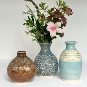 May include: Three ceramic vases of varying shapes and finishes. The left vase is round with a brown textured glaze. The center vase is blue with a white swirl pattern and holds a bouquet of green leaves and a pink flower. The right vase is light blue.