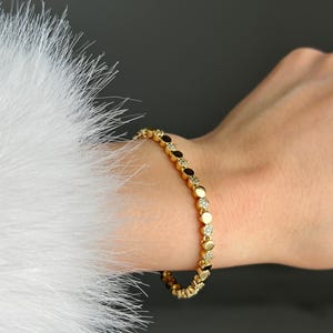 May include: Gold bracelet with alternating dark and clear stones. The bracelet is worn on a wrist, with a white, feathery texture in the background. The bracelet's design features small, round stones.