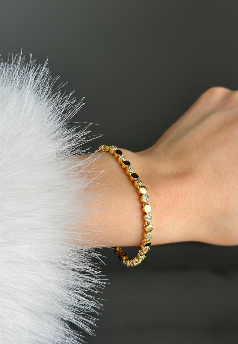May include: Gold bracelet with alternating dark and clear stones. The bracelet is worn on a wrist, with a white, feathery texture in the background. The bracelet's design features small, round stones.