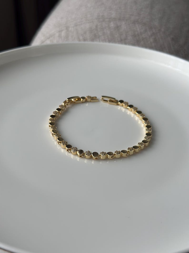 May include: A gold-coloured bracelet with a delicate design. The bracelet features small, round links, some adorned with sparkling accents. The bracelet is displayed on a white surface, showcasing its elegant simplicity. A classic piece of jewellery.