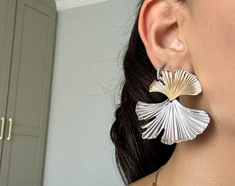 May include: Large, statement earrings in a fan-like design. The top portion is gold-toned, while the lower section is silver-toned. The earrings are worn on the ear, with a small, sparkly stud visible above the earring.