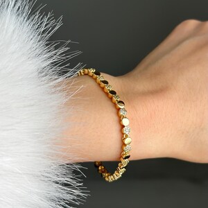 May include: Gold bracelet with circular and diamond-shaped links. The bracelet is worn on a wrist, with a white, feathery texture on the left. The background is dark grey. A stylish piece of jewellery.
