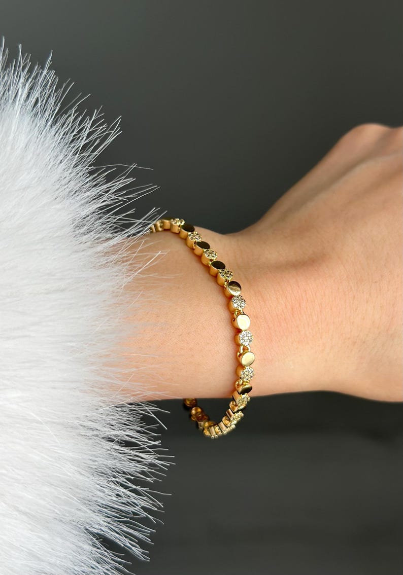 May include: Gold bracelet with circular and diamond-shaped links. The bracelet is worn on a wrist, with a white, feathery texture on the left. The background is dark grey. A stylish piece of jewellery.