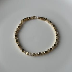 May include: A gold-coloured bracelet featuring a series of round, bezel-set clear crystals and gold-coloured metal links. The bracelet is displayed on a white surface. The clasp is open, showing the bracelet's construction.