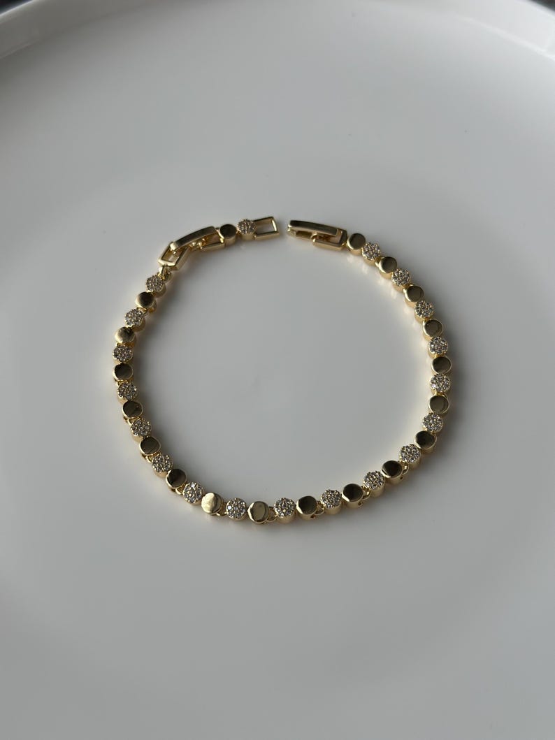 May include: A gold-coloured bracelet featuring a series of round, bezel-set clear crystals and gold-coloured metal links. The bracelet is displayed on a white surface. The clasp is open, showing the bracelet's construction.