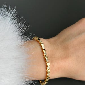 May include: A gold bracelet featuring a series of circular links, some set with small, sparkling stones. The bracelet is worn on a wrist, with a white, fluffy texture in the background.