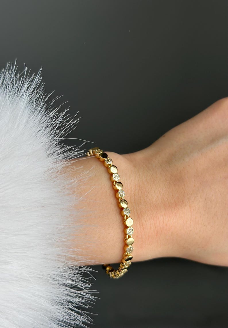 May include: A gold bracelet featuring a series of circular links, some set with small, sparkling stones. The bracelet is worn on a wrist, with a white, fluffy texture in the background.