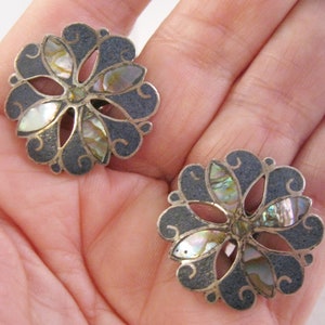 以下が含まれることがあります： 花柄のシルバーイヤリングのペア。イヤリングはスターリングシルバー製で、黒のエナメル地に真珠貝のアクセントが施されています。