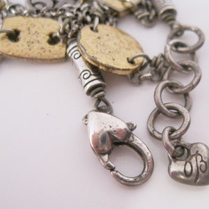 May include: A silver charm bracelet with a heart-shaped clasp and a charm that says "OB". The bracelet has several gold-toned charms, including a round charm with a design on it.
