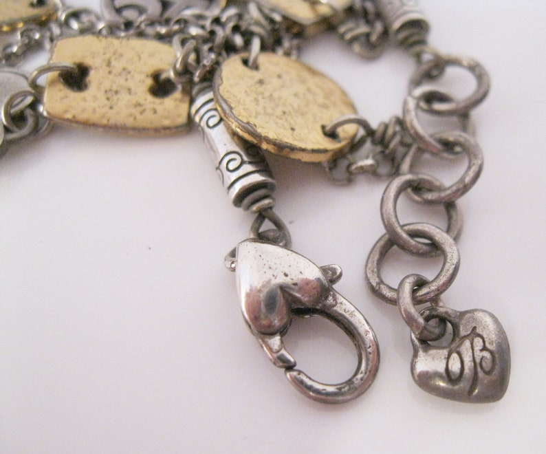 May include: A silver charm bracelet with a heart-shaped clasp and a charm that says "OB". The bracelet has several gold-toned charms, including a round charm with a design on it.