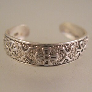 May include: A silver-colored toe ring with an open design. The ring features a repeating pattern of crosses and floral shapes, with a textured background. The ring is likely made of sterling silver.