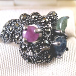 May include: A silver-toned ring with a floral design, featuring a central pink gemstone and two smaller blue and green gemstones. The ring is set with small, dark, faceted stones.