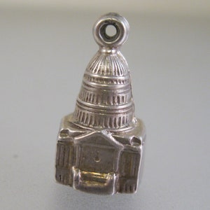 May include: Silver charm of the US Capitol building. The charm is detailed and features a dome and columns. It has a loop at the top for attaching to a bracelet or necklace.