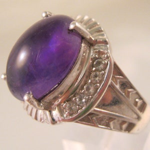 Könnte beinhalten: Ein silberner Ring mit einem großen, ovalen Amethyst, der in der Mitte eingesetzt ist. Der Ring ist mit kleineren, klaren Edelsteinen um den Amethyst herum verziert.