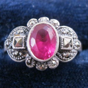 May include: A silver ring featuring a large, oval-cut, magenta-colored gemstone as the centerpiece. The ring is adorned with smaller, round and square marcasite stones, adding sparkle and detail to the design. The ring is set against a dark blue background.