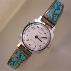 Op de afbeelding: Een zilverkleurig Timex horloge met een witte wijzerplaat en zwarte wijzers. De horlogeband is zilverkleurig met ingelegde turquoise gekleurde stenen. De wijzerplaat toont het woord "TIMEX" en "WATER RESISTANT".