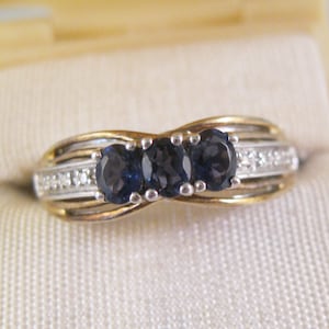 Könnte beinhalten: Ein Gold- und Silberring mit drei blauen Edelsteinen und einer Reihe kleiner Diamanten.
