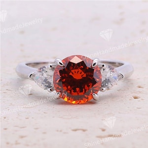 May include: A silver ring featuring a large, round, fiery orange gemstone as the centerpiece, flanked by two smaller, pear-shaped, clear gemstones. The ring is set against a neutral, textured background, highlighting the jewelry's sparkle and design.