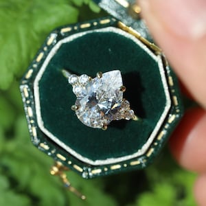 以下が含まれることがあります： 大きなペアシェイプのダイヤモンドを中央に、小さなダイヤモンドで囲んだゴールドリング。 金色のトリムが付いた濃い緑色のベルベットの箱に入っています。 リングのデザインはエレガントで、特別な機会に最適です。