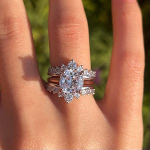 Könnte beinhalten: Ein dreiteiliges Ring-Set mit einem großen, klaren, kissenförmigen Mittelstein, umgeben von kleineren Akzentsteinen. Das Set enthält ein roségoldenes Band und zwei zusätzliche Bänder mit pavébesetzten Steinen. Die Ringe befinden sich an einem Finger.