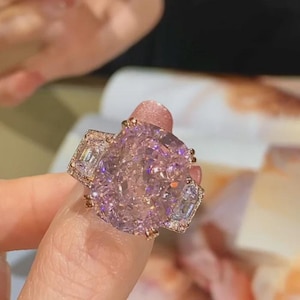 Puede incluir: Un anillo ornamentado con una gran piedra central cuadrada de color púrpura claro. El anillo tiene una banda dorada y está adornado con piedras preciosas rectangulares más pequeñas y transparentes a cada lado de la piedra principal. El anillo está engastado con múltiples garras.
