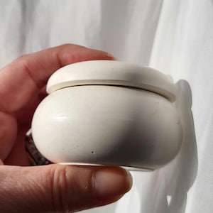 May include: A small, off-white ceramic jar with a matching lid. The jar is round with a slightly bulbous body and a flat, circular lid. The jar is held in a hand against a white background. The jar appears to be handmade.