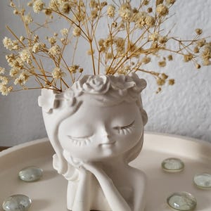 May include: A white ceramic face-shaped vase holding dried flowers. The vase is on a white tray with clear decorative stones. The face has closed eyes and a serene expression, with a floral crown.