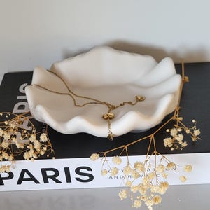 May include: A white, shell-shaped decorative dish holds a gold-coloured necklace. The dish sits atop a black book with the word "PARIS" in white. Dried flowers are arranged around the dish.