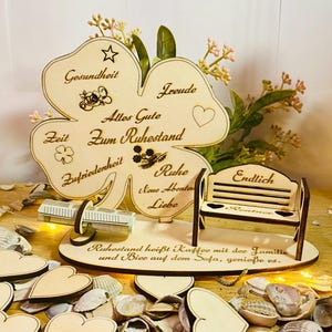 May include: A wooden four-leaf clover with the words "Gesundheit", "Freude", "Alles Gute Zum Ruhestand", "Zeit", "Zufriedenheit", "Ruhe", "Neue Abenteuer", "Liebe", and "Endlich" engraved on it. A small wooden bench is next to the clover. The clover is sitting on a surface covered in seashells.