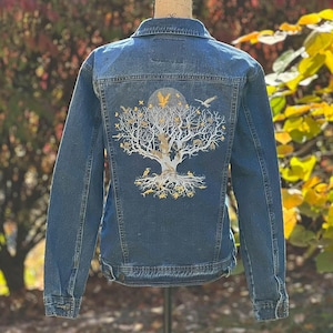 May include: A blue denim jacket with a white tree of life design on the back. The tree is adorned with gold birds, leaves, and other details.