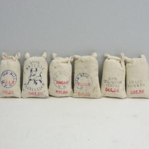 May include: Six small, off-white burlap sacks with red and blue text. The sacks are labeled with various items: Salt, Fertilizer, Sugar, Flour, Colombian Coffee, and Idaho Potato. Each sack is marked with "50 LBS".