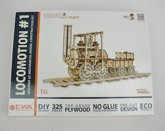 May include: A white rectangular box for a Locomotion #1 wooden 3D mechanical model construction kit. The box features an image of a light brown wooden steam locomotive on tracks, with text including 325 pieces, 1:25 scale, and DIY assembly.