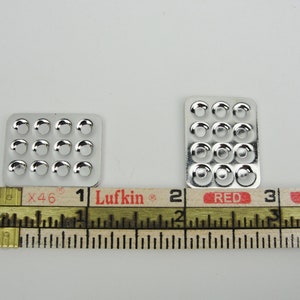 May include: Two silver metal trays with circular indentations. The trays are 1 inch and 2 inches wide, respectively. The trays are measured with a ruler labeled "Lufkin" and "Red".