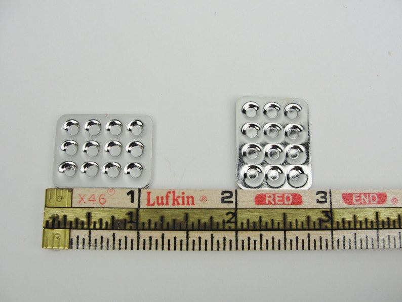 May include: Two silver metal trays with circular indentations. The trays are 1 inch and 2 inches wide, respectively. The trays are measured with a ruler labeled "Lufkin" and "Red".