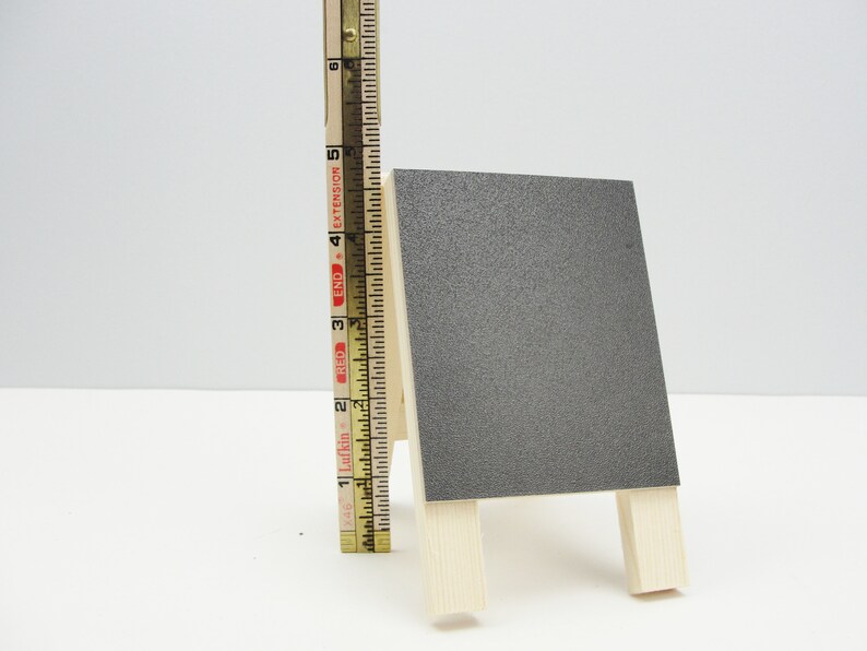 May include: A small, gray chalkboard easel with a wooden frame. The easel is about 6 inches tall.