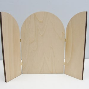 May include: A three-panel wooden screen with a light wood finish and dark brown edges. The center panel has a rounded top, and all panels are connected by brass hinges. The screen is open, displaying its full width.