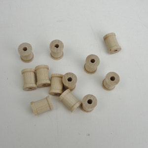 May include: A group of ten wooden spools, all the same size and shape, are scattered on a white surface. The spools are light brown in color and have a hole in the center.